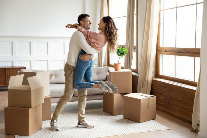 Een pas verhuisd jong koppel staat in elkaars armen in hun woonkamer vol kartonnen dozen
